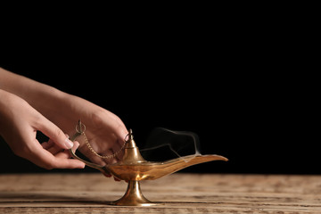 Woman rubbing magical Aladdin lamp on table against dark background