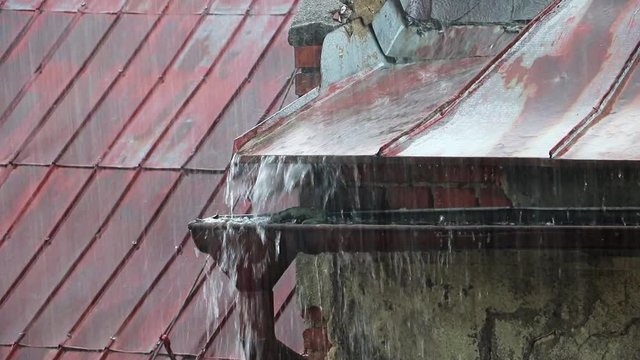 A heavy rain fall on a metal roof. The stream of rainwater flows into the full eaves.