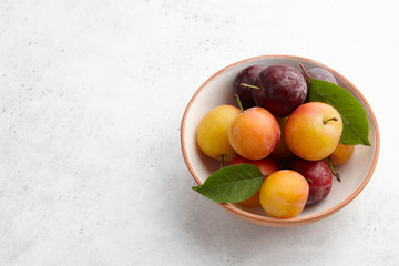 Fresh colorful plums with leaves in fruit bowl on white stone background, seasonal food