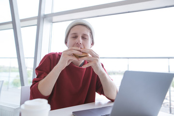 Young man in a casual dress sits with a notebook in a fast-food restaurant and eats a burger. Student bites sandwich. Work and meals at the fast food cafe