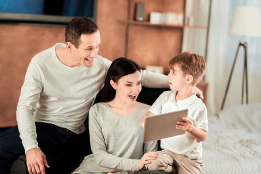 Look here. Pleasant young boy holding a tablet while showing a picture to his parents