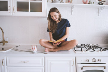Young girl reading an interesting book in the kitchen. Woman drinking morning coffee. Before the...