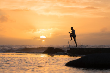 Fototapeta na wymiar Silhouette of Man Leaping Off Sea Wall at Sunset with Copy Space