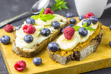 Traditional American and European summer breakfast: sandwiches of toast with peanut butter or cream cheese with raspberries and blueberry, banana. Black concrete table. copy space top view