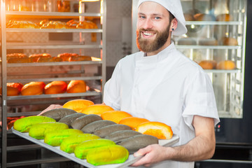 A young handsome baker is holding colorful fresh baked goods on a sheet on the background of the oven