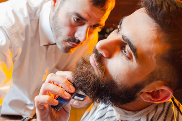 Barber cuts a beard with a trimmer to a young handsome guy with a mustache. Haircut of a beard in a male barber shop