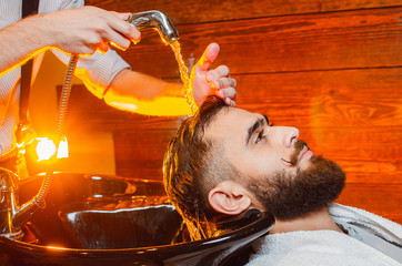 Barber washes the head of a young handsome guy with a beard and mustache in the sink. Male beauty salon