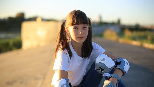 girl on rollers in a white t-shirt and two pigtails sitting on the curb on the side of the road and watching the sunset. large portraits of Caucasian girls.