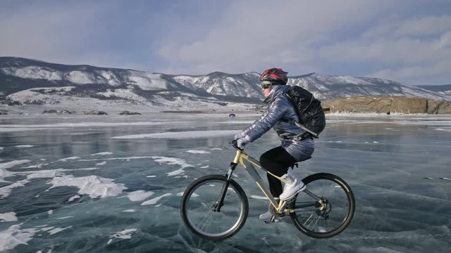Woman is riding bicycle on the ice. The girl is dressed in a silvery down jacket, cycling backpack and helmet. Ice of the frozen Lake Baikal. The tires on the bicycle are covered with special spikes
