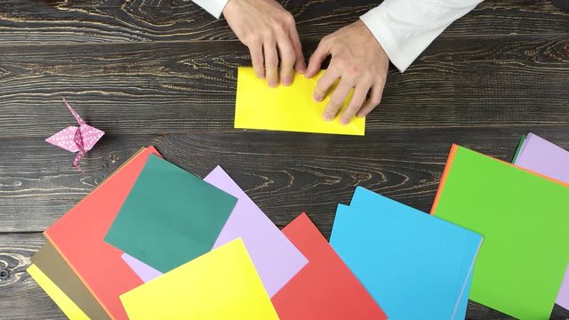 Male hands making origami with yellow paper. Colourful papers on wooden table, top view.
