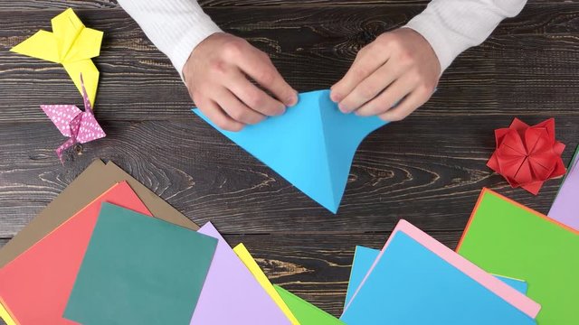 Man folding origami figure from blue paper. Male hands creating origami form, top view.