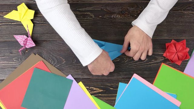 Male hands creating origami with blue paper, top view. Wooden desk surface background.