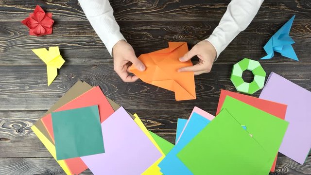 Hands making origami figure with orange paper. Top view. Wooden desk surface.