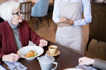 Senior woman sitting at restaurant with salad on the table and making an order for her neighbor