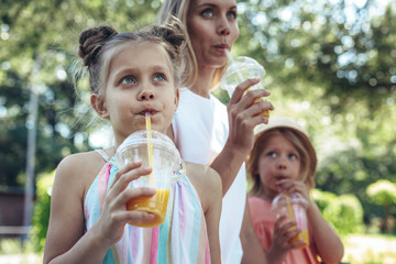 Waist up portrait of small girl sitting outside beside her mother and sister and sipping juice...