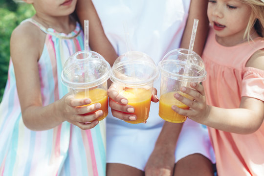 Cheers. Close up of female hands holding plastic cups with orange juice. Mother and kids are clinking glasses in park enjoying summer together