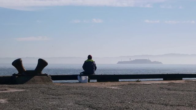 A lone fisherman casts from the pier at Crissey Field on a sunny spring morning.  Alcatraz visible in the distance.  Fisherman's back is to the camera.  Gravel surface of the pier in the foreground.