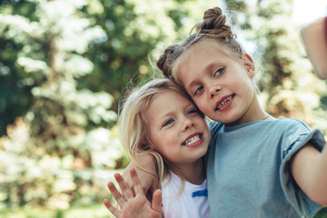 Waist up portrait of small girls standing and looking at mobile while making selfie. They are bonding to each other with smile and delight