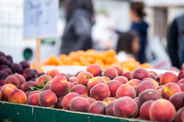 Bin filled with fresh peaches at a farmers market.  Shoppers and signage out of focus in the background.