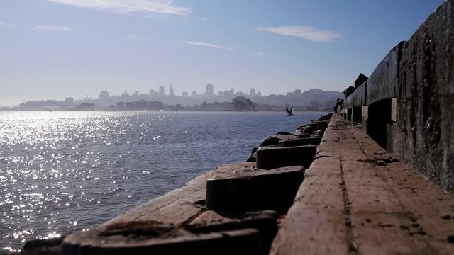 A view of downtown San Francisco taken from the fishing pier at Crissey Field.  Creosote coated pier timbers, fisherman sitting at the far end.  Bright morning sun sparkles on the bay. 