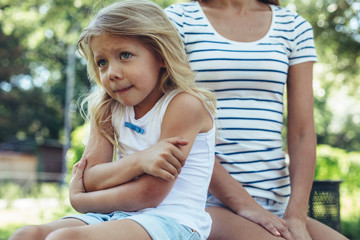 Waist up portrait of resented girl sitting in park with turned back beside adult. She is folding hands and looking angry