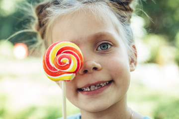 Waist up portrait of small smiling young girl holding lollypop outdoor. She is covering eye with candy and laughing with joy