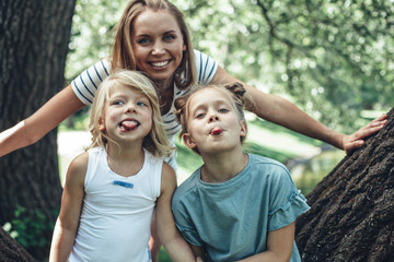 Waist up portrait of smiling mother and daughters standing by tree and showing tongues. They content and happy to rest on nature