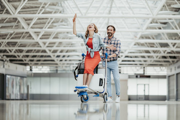Fototapeta premium Full length portrait of laughing girl and cheerful bearded male having fun in airport. Entertainment of happy couple concept
