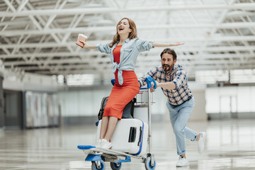 Full length portrait of beaming woman gesticulating hands while riding on luggage trolley. Laughing...