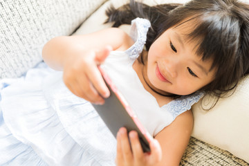 Obraz na płótnie Canvas Cute little girl having fun to play game on smart phone lying on sofa.