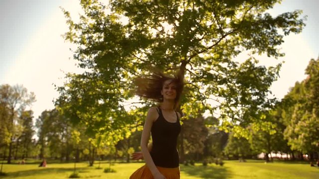Attractive hzandsome pretty young female with long wavy hair is walking in park in summer day, turning to a camera. Steadicam smooth camera movement. Slow motion shot
