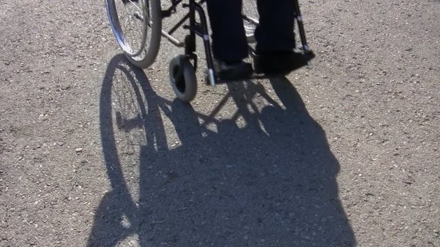 On a sunny day man, wheelchair user and his shadow is coming towards the camera on plain asphalt background 