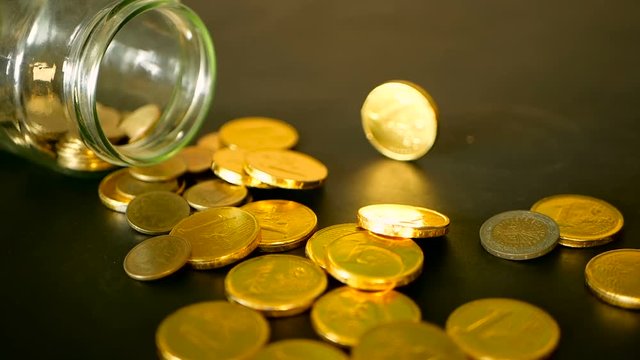 Yellow coins fell out from jar. Symbol of investing, keeping money concept. Collecting cash conis in glass tin as moneybox. Close-up still life with gold coins on black table and rotating penny.
