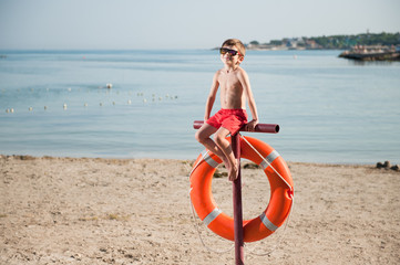 Fototapeta na wymiar healthy little boy in sunglasses sitting on beach with lifebuoy in summer day