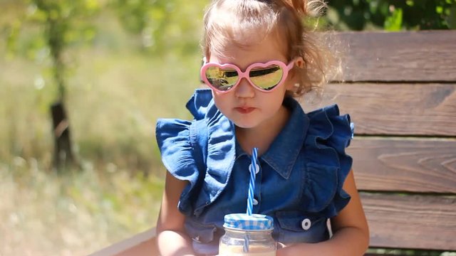 Child girl in sunglasses drinks smoothies in the park on a sunny windy day