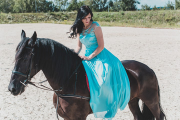 Beautiful young woman in long blue dress riding a horse in countryside. Portrait of a dark horse and woman. Attractive girl riding on horse rural location 