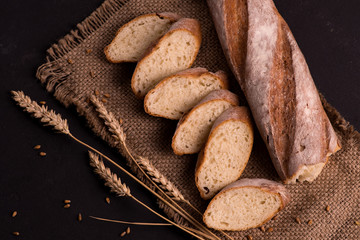 Pieces of sliced ​​baguette and half baguette lie on a dark jute napkin.  Nearby lie a few spikelets of wheat