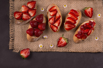 Five pieces of baguette with sliced ​​strawberries lie on a jute napkin on a dark table. On a napkin lie white, pink flowers