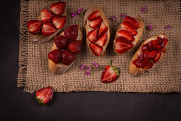 Five pieces of baguette with sliced ​​strawberries lie on a jute napkin on a dark table.
