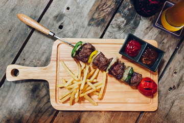 BBQ beef kebab served with French fries, tomato sauce and BBQ sauce on wooden plate.