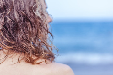 Curly haired brown-haired girl standing on the beach with her back, hair care at sea concept