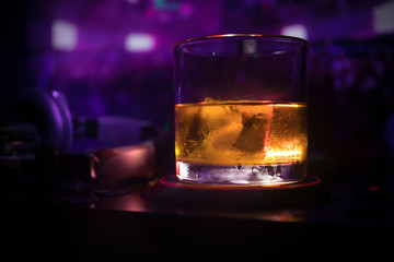 Glass with whisky with ice cube inside on dj controller at nightclub. Dj Console with club drink at music party in nightclub with disco lights.