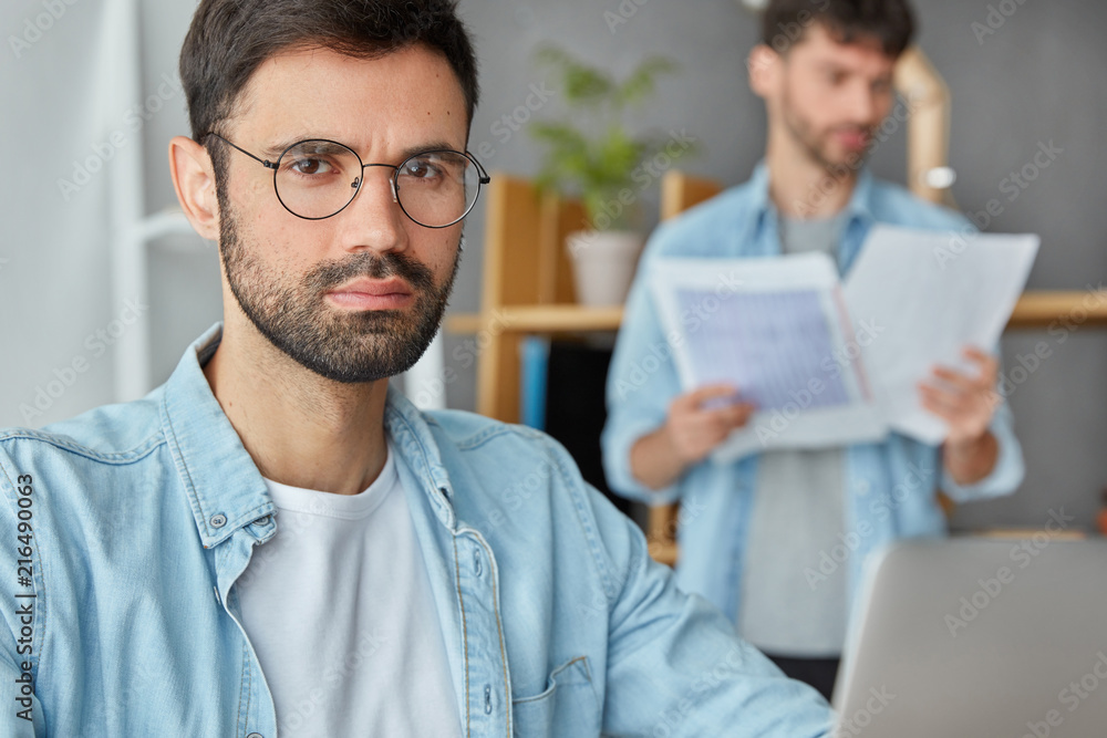 Wall mural Serious bearded male entreprenuer works at business project on laptop, looks confidently at camera, his colleague stands in background, studies financial papers, pose at meeting room at office