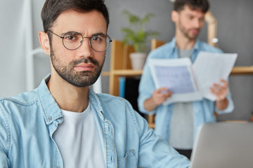 Serious bearded male entreprenuer works at business project on laptop, looks confidently at camera, his colleague stands in background, studies financial papers, pose at meeting room at office