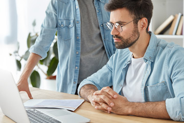 Indoor shot of two men discuss together work questions, make financial report on laptop computer, work with documents. Male students prepare common project. Cooperation and business concept.