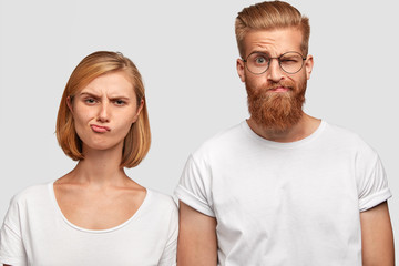 Obraz premium Discontent female and male coworkers purse lips and frown faces, dislike their plan to improve financial situation, wear casual t shirts, stand next to each other, isolated over white background