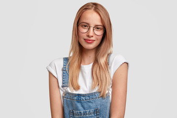 Waist up portrait of pretty young European female with long hair, wears glasses and overalls, feels satisfied to hear results of exam, looks with friendly expression, isolated over white background