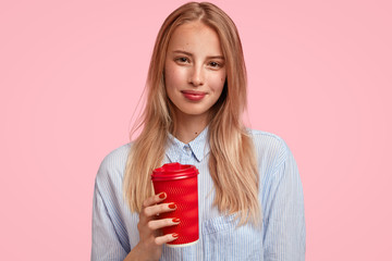 Attractive blonde female student holds hot beverage in disposable paper cup, wears elegant shirt, stands against pink background, has break after having lectures. People and spare time concept