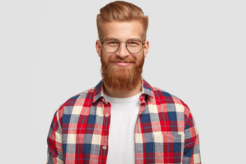 Glad ginger male with pleased expression, wears glasses and fashionable checkered shirt, rejoices...