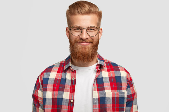 Glad ginger male with pleased expression, wears glasses and fashionable checkered shirt, rejoices successfully made project, poses alone against white background. People, emotions, lifestyle